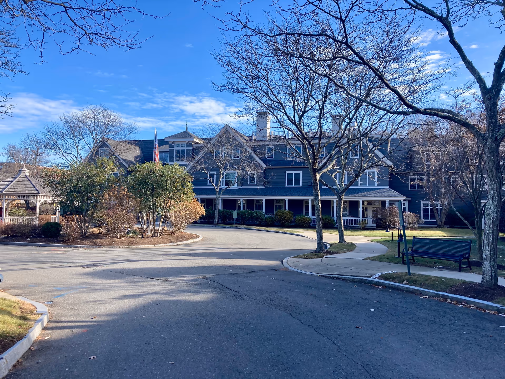 Exterior view of a large, multi-story assisted living facility building with a dark blue facade and white trim. The building is surrounded by leafless trees, bushes, and a paved driveway leading to the entrance. There is a gazebo on the left side and a bench on the right side near a sidewalk. The sky is clear with some clouds.