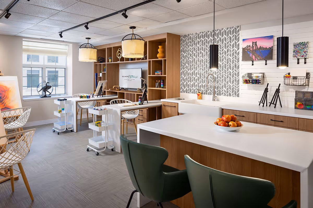 A bright, modern activity room in a senior living facility featuring a large white countertop island with green chairs, a bowl of apples, a sink, and wooden cabinetry. The back wall has patterned wallpaper, hanging black pendant lights, and art supplies organized on shelves and in baskets. There are two white rolling carts with art materials, wire chairs, and a window letting in natural light. A TV screen displays the Belmont Village Senior Living logo.