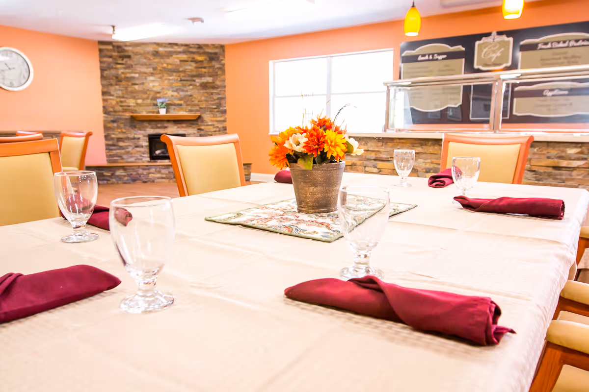 A dining room table set with a white tablecloth, clear drinking glasses, and burgundy cloth napkins. A centerpiece with orange and white flowers in a metal pot is placed in the middle of the table. The room has orange walls, a stone fireplace, and a serving counter with menu boards in the background.