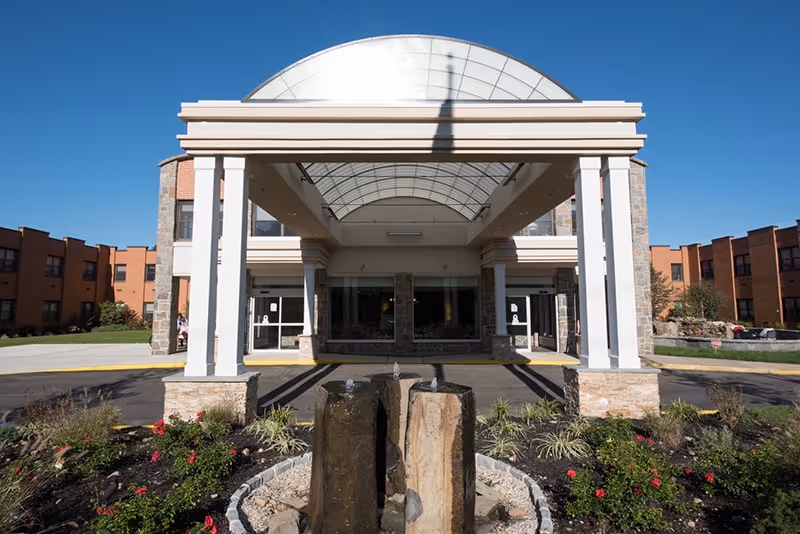 Front exterior view of St. James Rehabilitation & Healthcare Center showing a covered entrance with white columns, a glass canopy, and a stone water fountain in the landscaped area in front of the building under a clear blue sky.