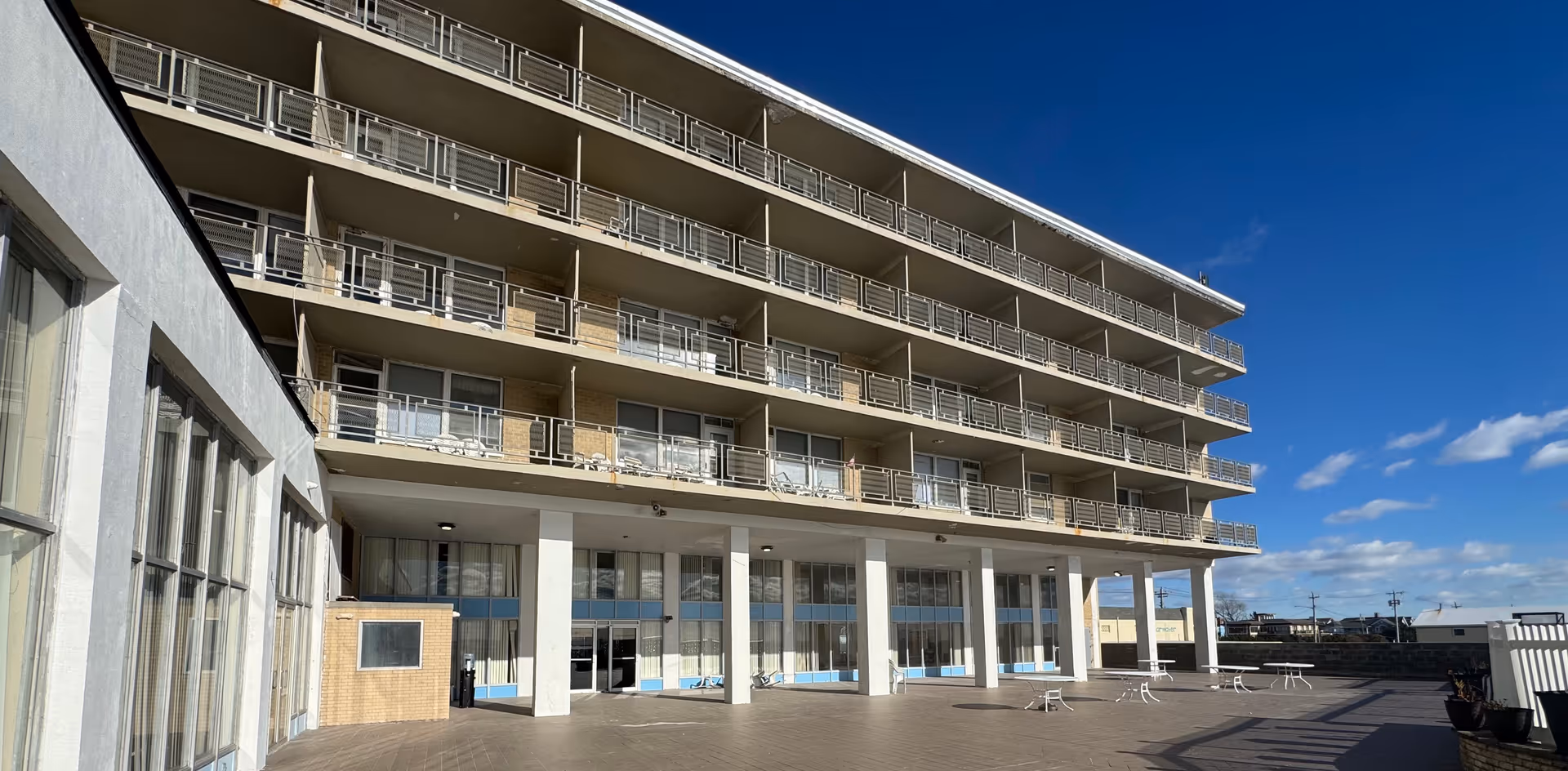 Exterior view of a multi-story building with balconies on each floor under a clear blue sky. The building has large windows on the ground floor and a spacious paved area in front with some outdoor furniture.