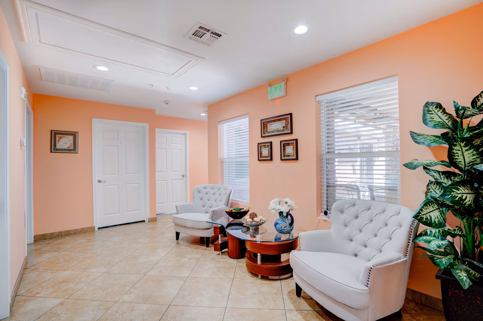 A cozy seating area in a senior living facility with two white tufted armchairs, a round wooden coffee table with decorative items including a vase with white flowers, and a large green potted plant. The walls are painted peach, and there are two windows with blinds and framed certificates or artwork on the wall. Two white doors are visible in the background.