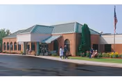 Exterior view of a single-story brick building with a green metal roof and an entrance canopy. Several people are sitting on benches near the entrance, and an American flag is visible on a flagpole to the right. The area is landscaped with grass and flowers.