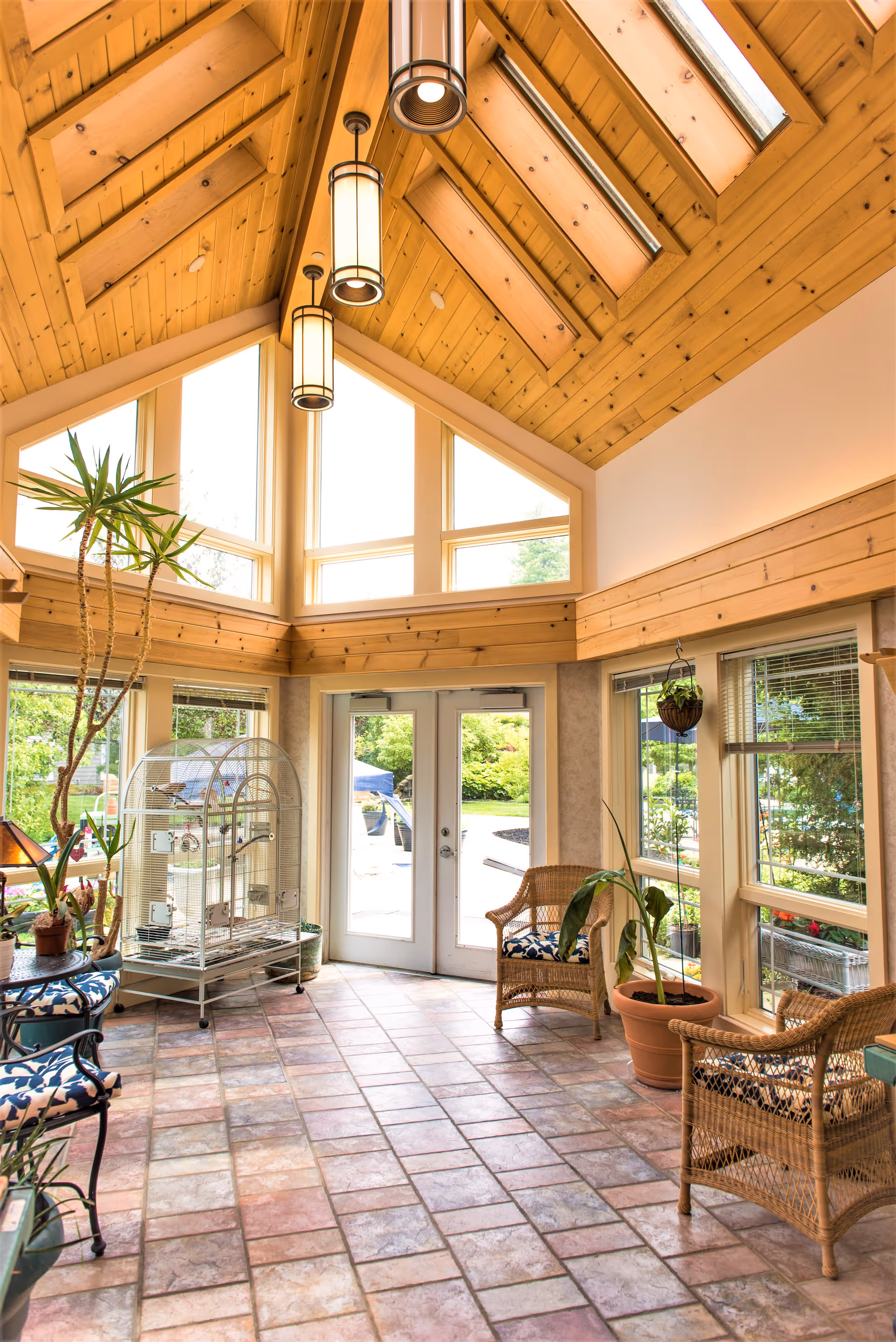 A bright sunroom with a high wooden ceiling featuring skylights and hanging pendant lights. The room has large windows and glass double doors leading outside. There are wicker chairs with patterned cushions, potted plants, and a birdcage inside the room. The floor is tiled with multicolored square tiles.