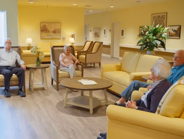 A cozy common area in a senior living facility with four elderly people sitting on yellow and beige armchairs and sofas arranged around a wooden coffee table. The room has light yellow walls, framed artwork, a potted plant, and soft lighting from table lamps.
