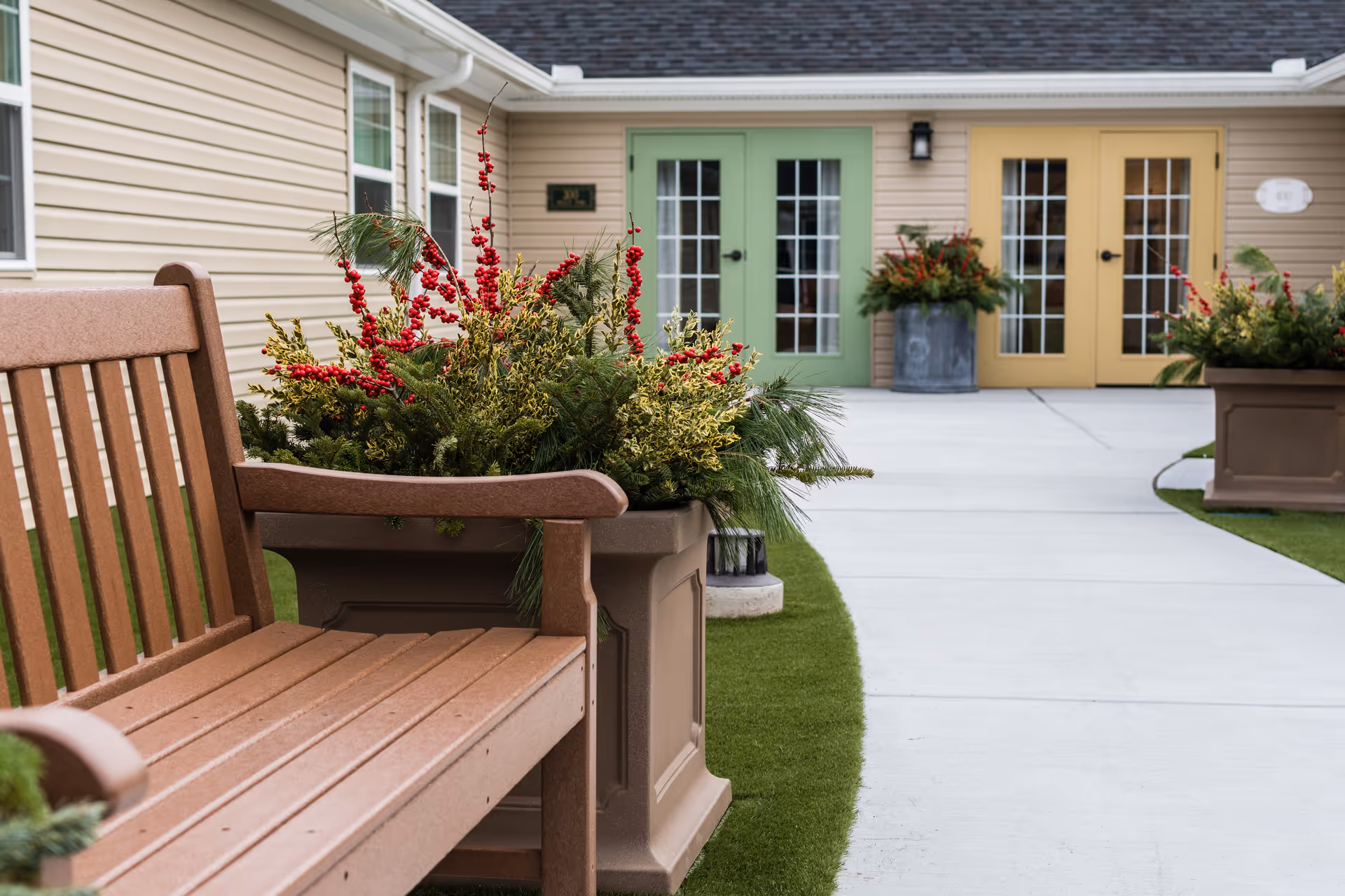 Outdoor seating area at a senior living facility featuring a wooden bench next to large planters filled with greenery and red berries. The background shows a beige building with two sets of French doors, one painted green and the other yellow, with additional planters near the entrances.