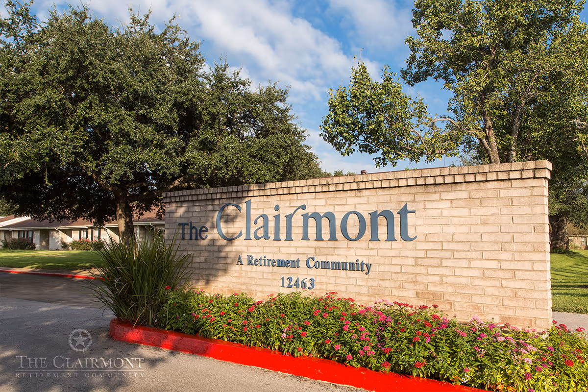 Brick sign at the entrance of The Clairmont Retirement Community with the address 12463, surrounded by green bushes and flowers, with trees and a partly cloudy sky in the background.