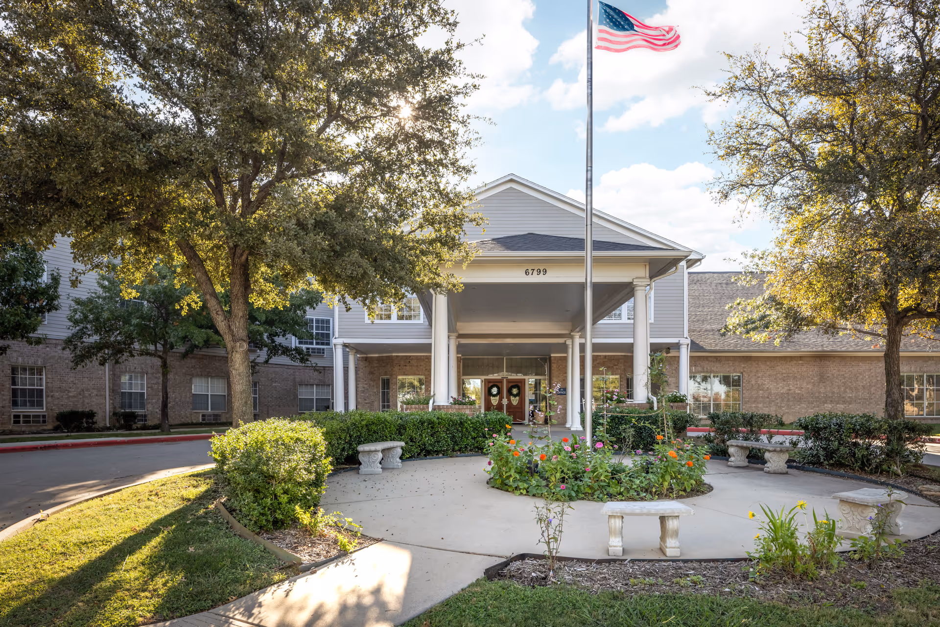 Front exterior view of The Waterford at Fort Worth facility with a circular driveway, a flagpole with an American flag, benches around a flower bed, and trees on either side.