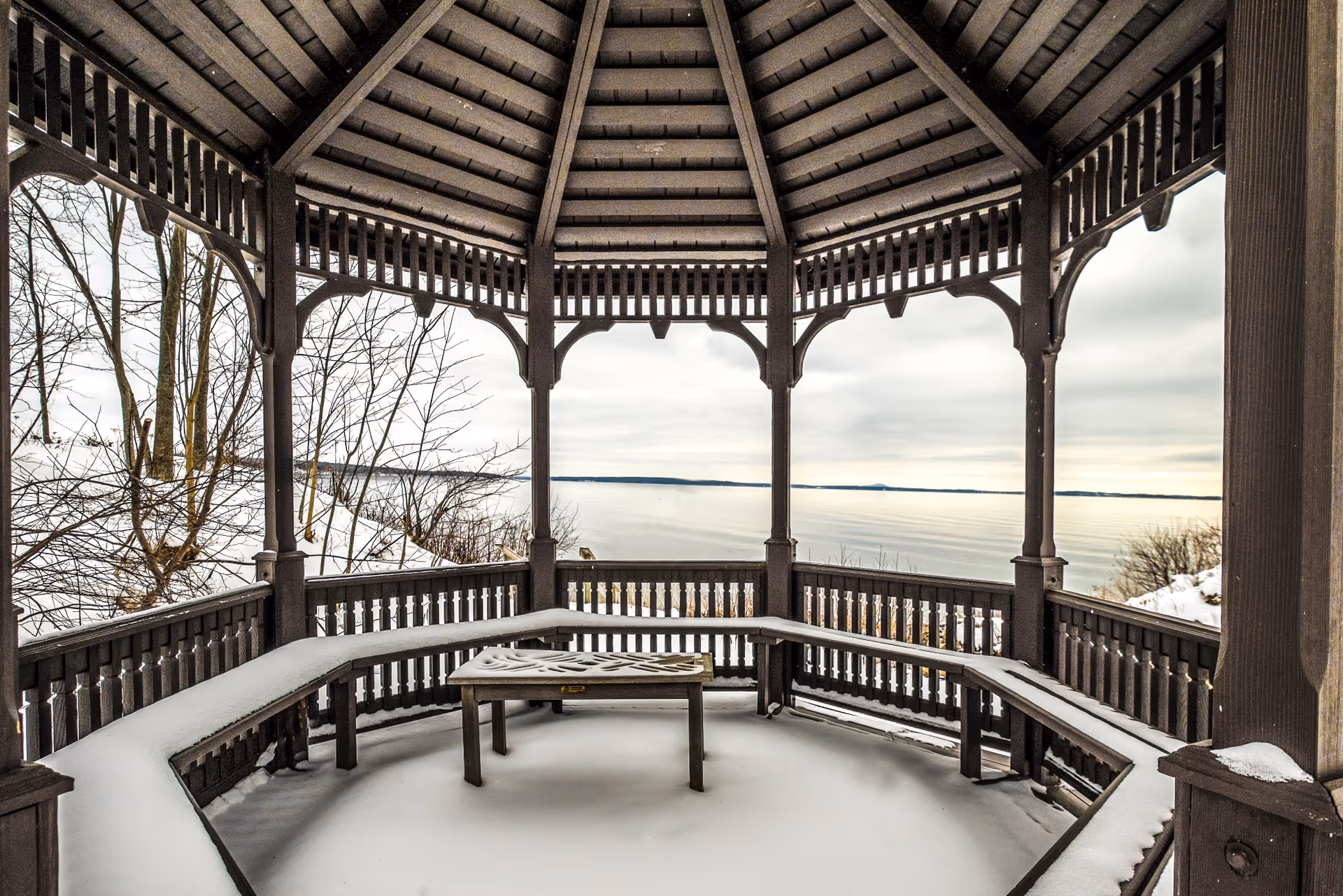A wooden gazebo with a snow-covered bench and table inside, overlooking a calm body of water with a snowy landscape and bare trees in the background under a cloudy sky.