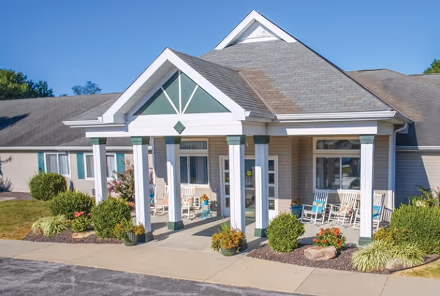 Front exterior view of a single-story senior living facility building with a covered entrance supported by white columns. There are rocking chairs on the porch and landscaped bushes and flowers around the entrance. The sky is clear and blue.