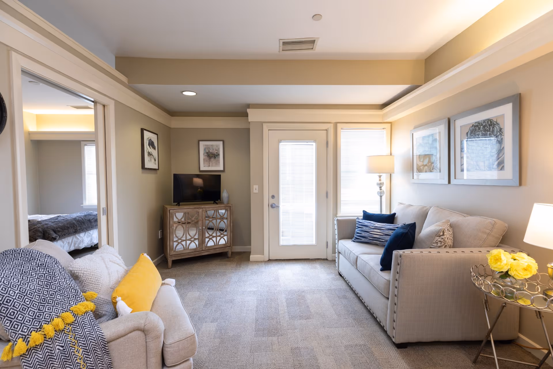 A cozy living room with two beige sofas adorned with blue and yellow pillows. A small mirrored cabinet with a TV on top is against the far wall, next to a door with blinds. There are two framed artworks on the wall above one sofa and a side table with a lamp and yellow flowers. An adjacent room with a bed is visible through an open doorway.