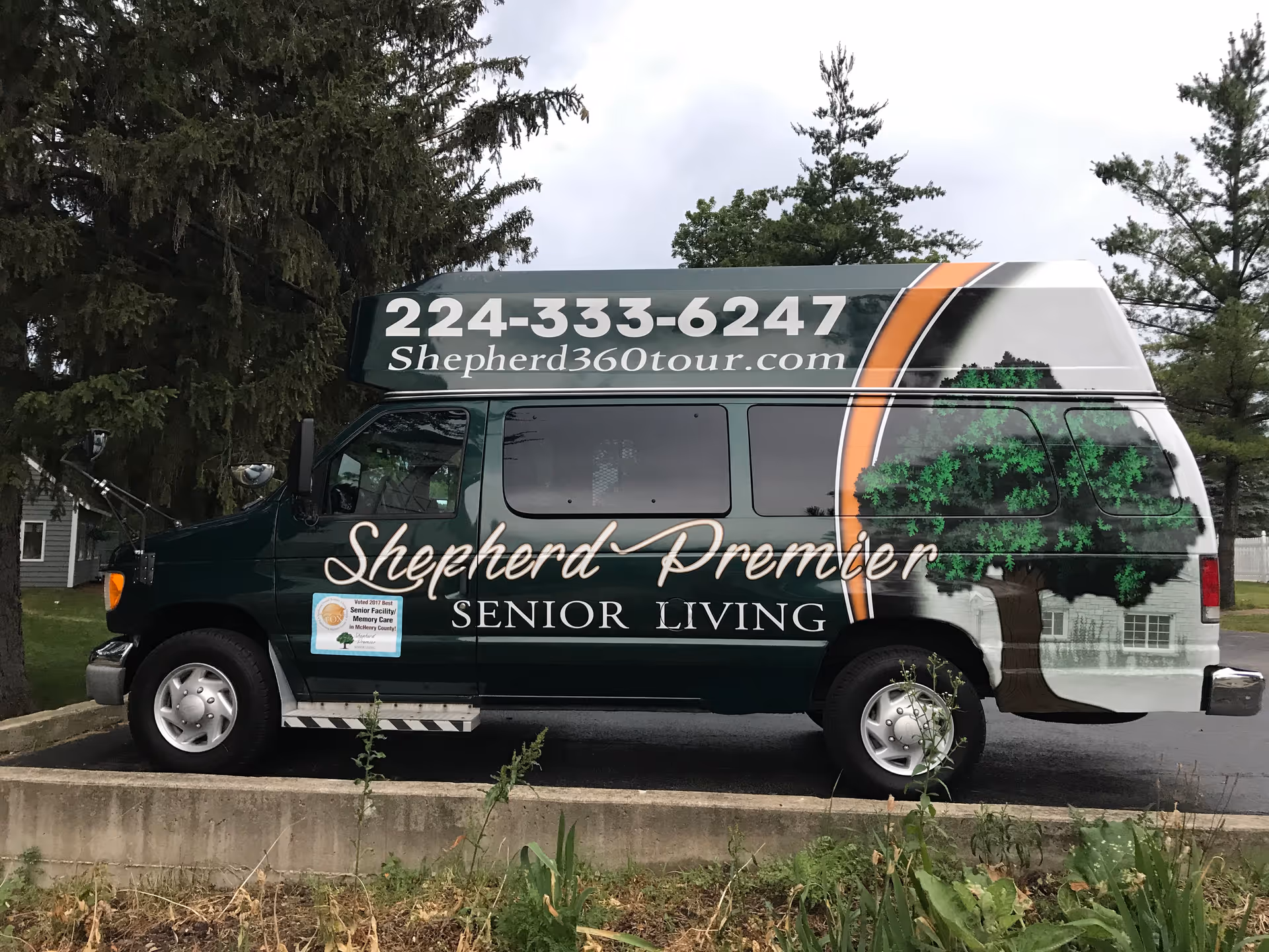 A dark green shuttle van parked outdoors with the branding 'Shepherd Premier Senior Living' on the side. The van features a large tree graphic and contact information including a phone number and website URL. The background includes trees and a cloudy sky.