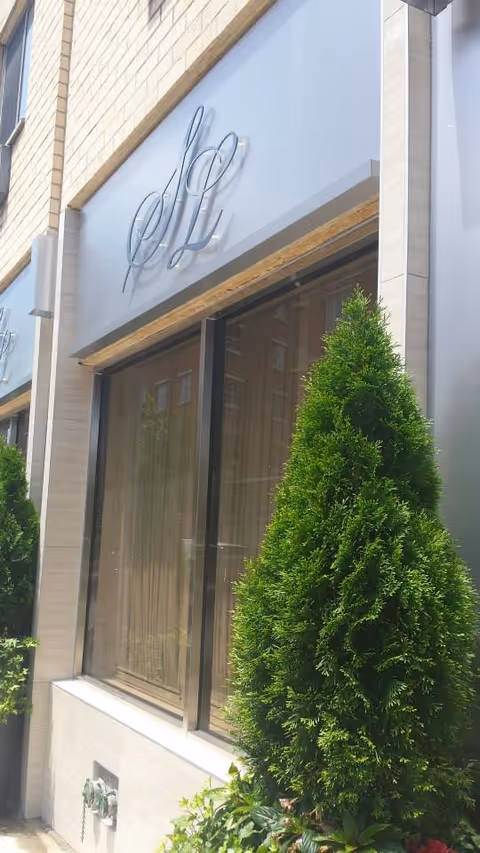 Exterior view of a building with large windows and a sign featuring the initials 'SL'. There are tall green shrubs planted in front of the building.