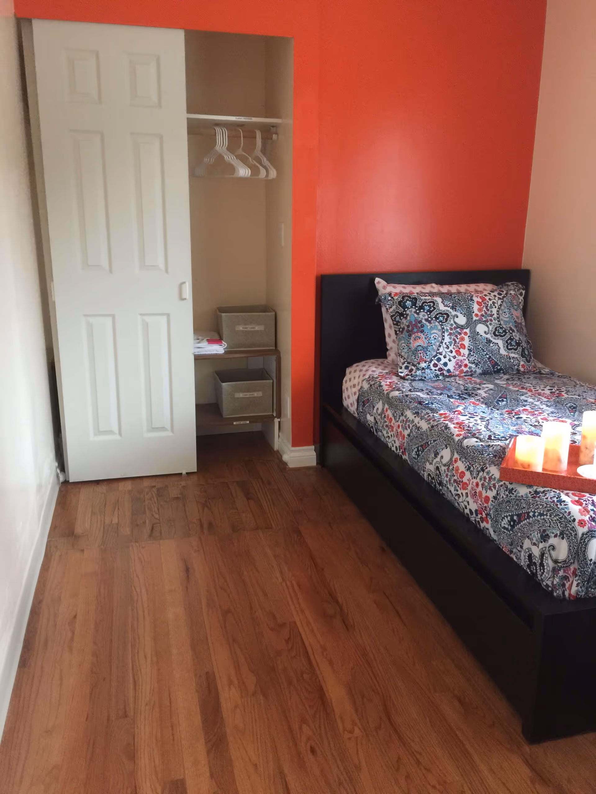 A small bedroom with a single bed featuring a colorful patterned bedspread and pillow. The bed has a dark frame and a wooden tray with candles on it. The room has hardwood flooring and an open closet with a sliding white door, containing several white hangers and two labeled storage bins on shelves. The wall behind the bed is painted bright orange, while the adjacent walls are light-colored.