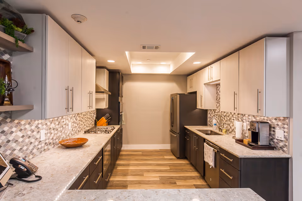 A modern kitchen with light-colored upper cabinets and dark lower cabinets, a marble countertop, a mosaic tile backsplash, a stainless steel refrigerator, a built-in oven, a gas stove, a sink with a faucet, a coffee maker, and wooden flooring. The kitchen is well-lit with recessed lighting and a skylight.