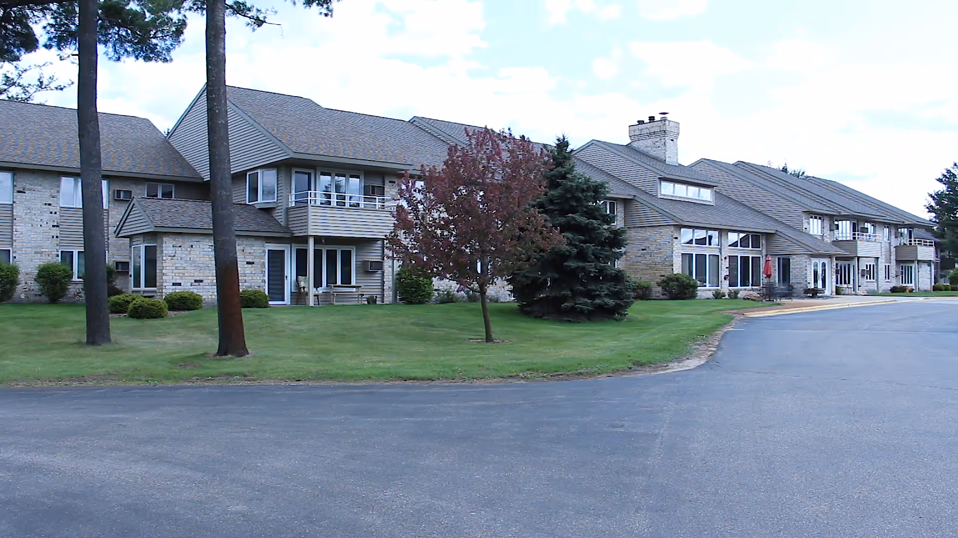 Exterior view of a two-story assisted living facility building with beige siding and stone accents, surrounded by green grass, trees, and a paved driveway under a partly cloudy sky.