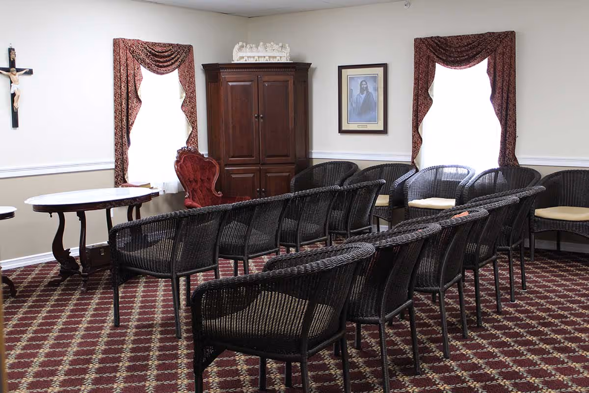 A small meeting or chapel room with rows of black wicker chairs facing a red upholstered armchair and a wooden cabinet. The room has two windows with patterned red curtains, a crucifix on the wall, a framed picture of Jesus, and a patterned carpet.