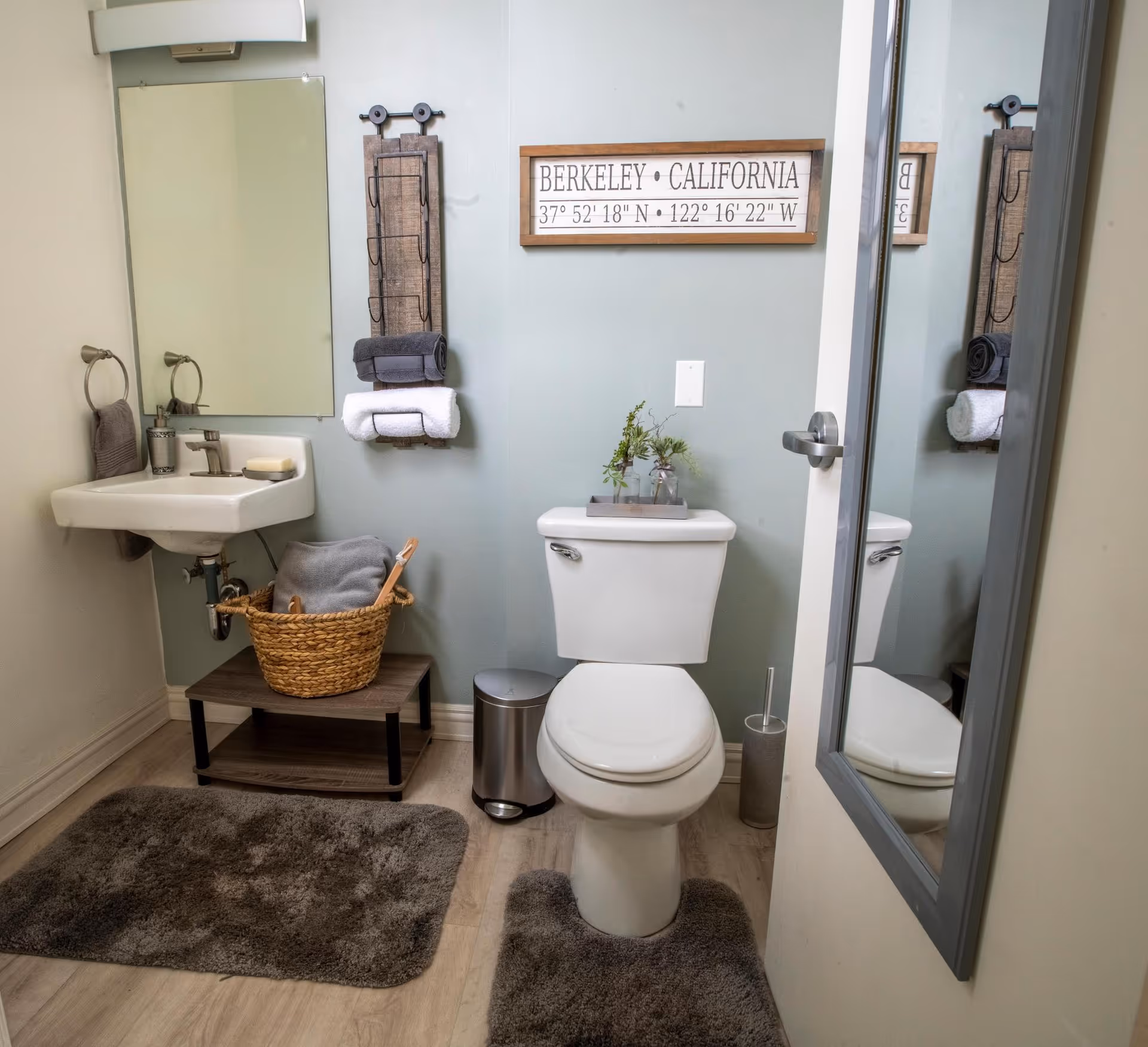 A small bathroom with a white toilet and a wall-mounted sink. There is a gray rug in front of the toilet and another in front of the sink. A wicker basket with towels sits on a small wooden shelf under the sink. Above the toilet is a decorative sign with the text 'BERKELEY • CALIFORNIA' and coordinates. A wall-mounted towel rack holds rolled towels, and a mirror is mounted on the door reflecting part of the bathroom.