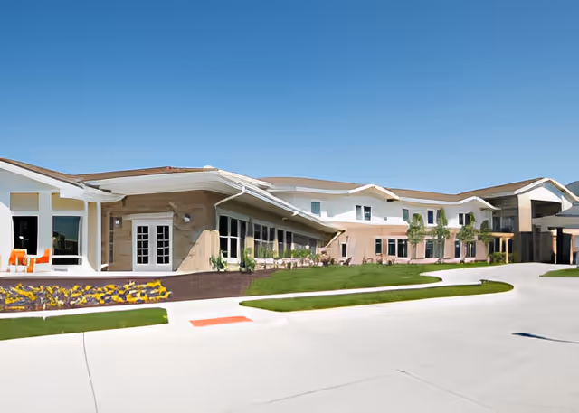 Exterior view of The Heritage at Sterling Ridge facility showing a large, modern building with beige walls and a brown roof under a clear blue sky. There is a well-maintained lawn and flower bed in front, along with a paved driveway and sidewalk.