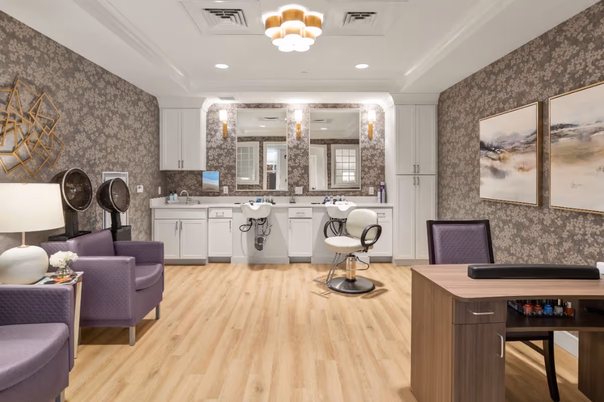A well-lit salon area in a senior living facility featuring two purple armchairs with a lamp and small table, two hair drying stations, a white salon chair in front of two sinks and mirrors, white cabinetry, a wooden desk with nail polish bottles, and decorative wall art on floral wallpaper.