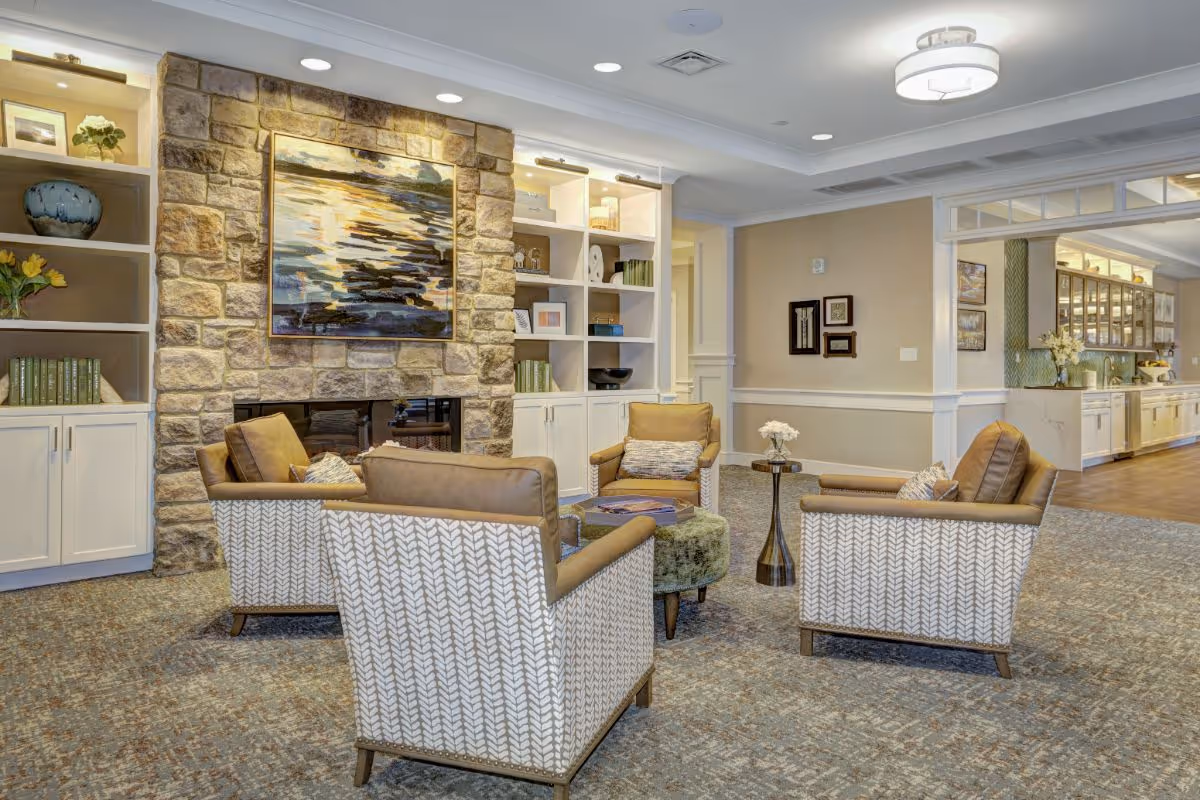 A cozy living room area with four patterned armchairs arranged around a small round ottoman and a side table with a flower vase. The room features a stone fireplace with a large abstract painting above it, built-in white shelves with decorative items, and a view into a kitchen area with white cabinetry and a wooden floor.