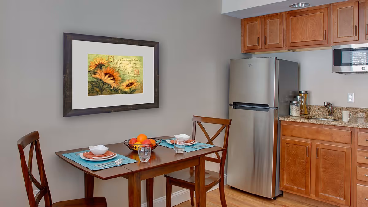 A small kitchen and dining area with wooden cabinets, a stainless steel refrigerator, a microwave, and a granite countertop. A wooden dining table set for two with plates, bowls, glasses, and a fruit basket is placed against a light gray wall with a framed sunflower painting.