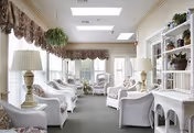 A bright and airy living room with white wicker furniture including armchairs and shelves. The room has large windows with floral valances, two table lamps with white shades, hanging plants, and skylights allowing natural light to fill the space.