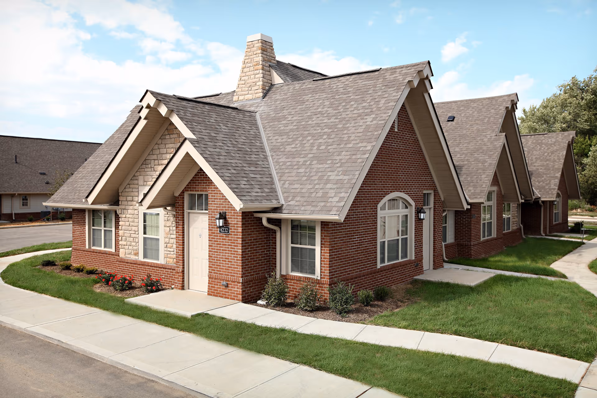 Exterior view of a brick and stone residential building with multiple peaked roofs and several windows, surrounded by green grass and sidewalks under a partly cloudy sky.