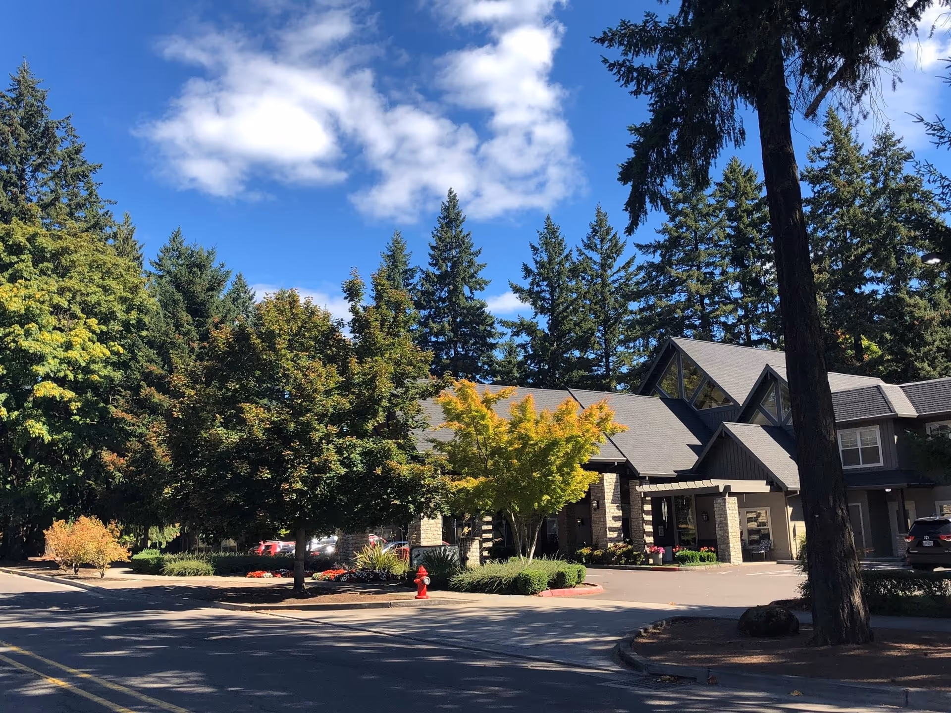 Exterior view of a senior living facility building surrounded by tall evergreen trees and other greenery under a blue sky with some clouds. The building has a sloped roof and stone accents, with a driveway and a red fire hydrant visible in front.