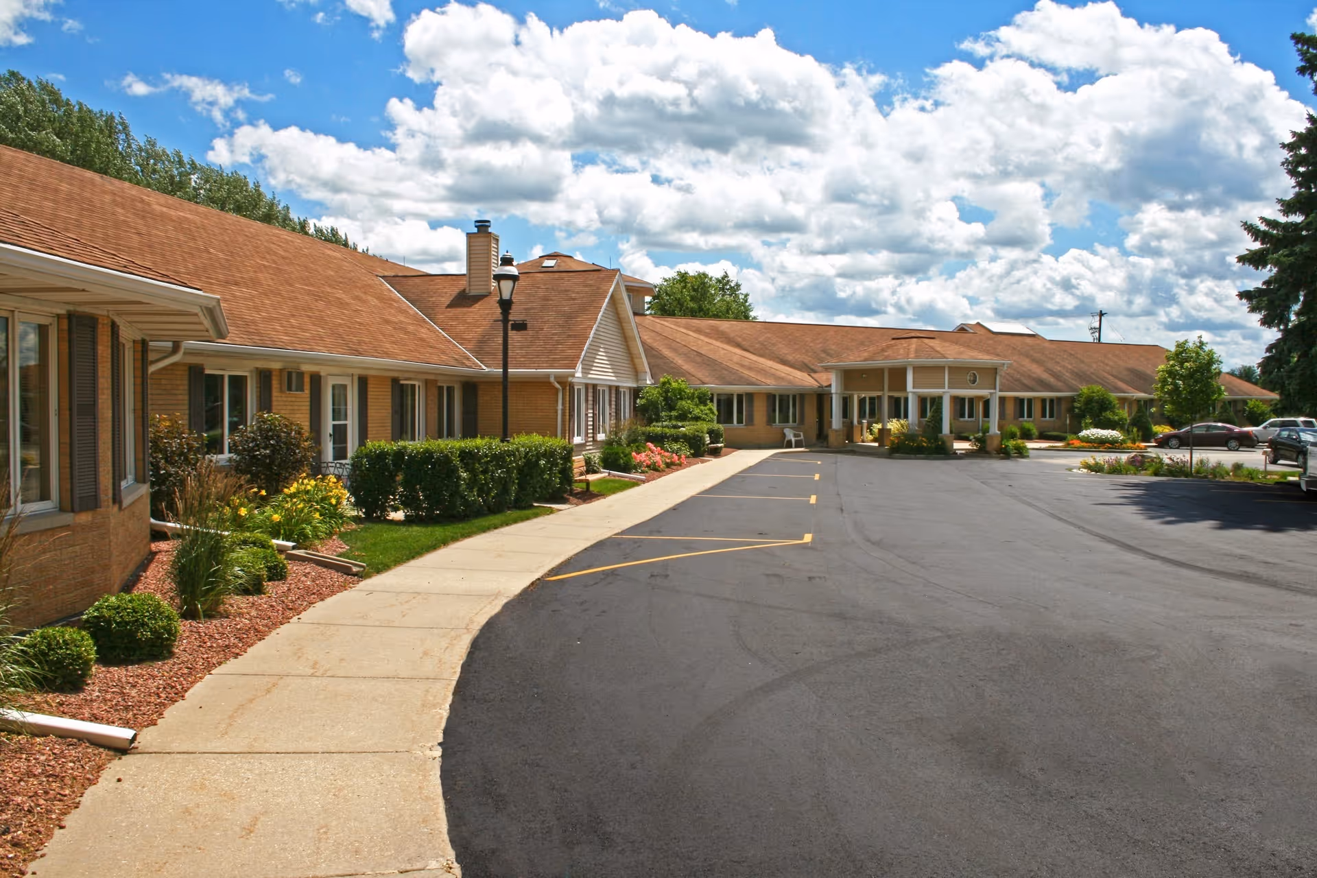 Exterior view of a single-story senior living facility with a long building featuring a brown roof and beige walls. There is a paved driveway and sidewalk with landscaped bushes and flowers along the building. The sky is partly cloudy with blue patches.