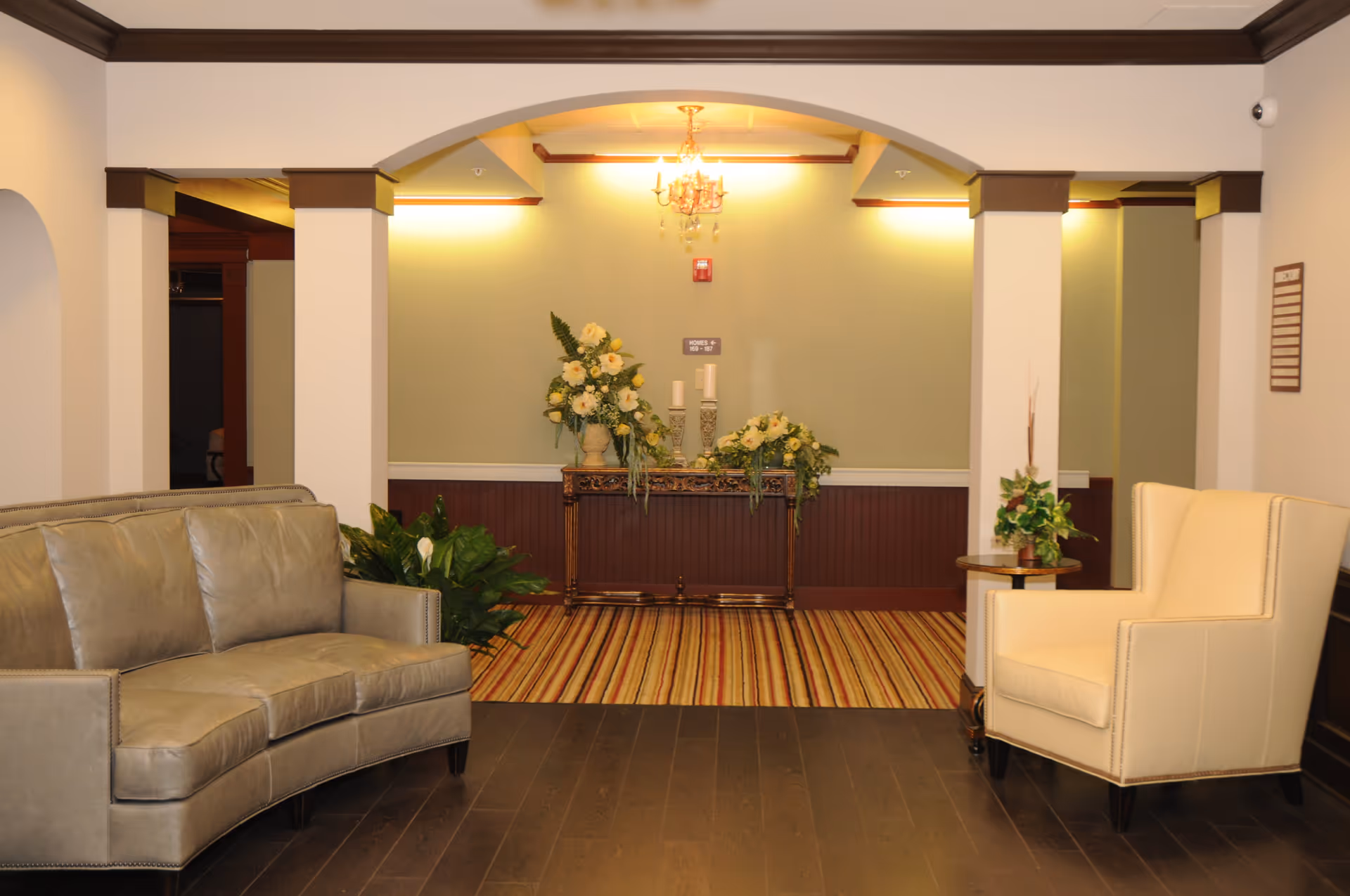 A cozy seating area in a senior living facility with a curved gray leather sofa on the left and a cream-colored armchair on the right. Between them is a small round wooden table with a plant. In the background, there is an archway leading to a hallway with a decorative wooden console table adorned with floral arrangements and candles. The floor is dark wood, and the walls are light with dark wood trim and crown molding.