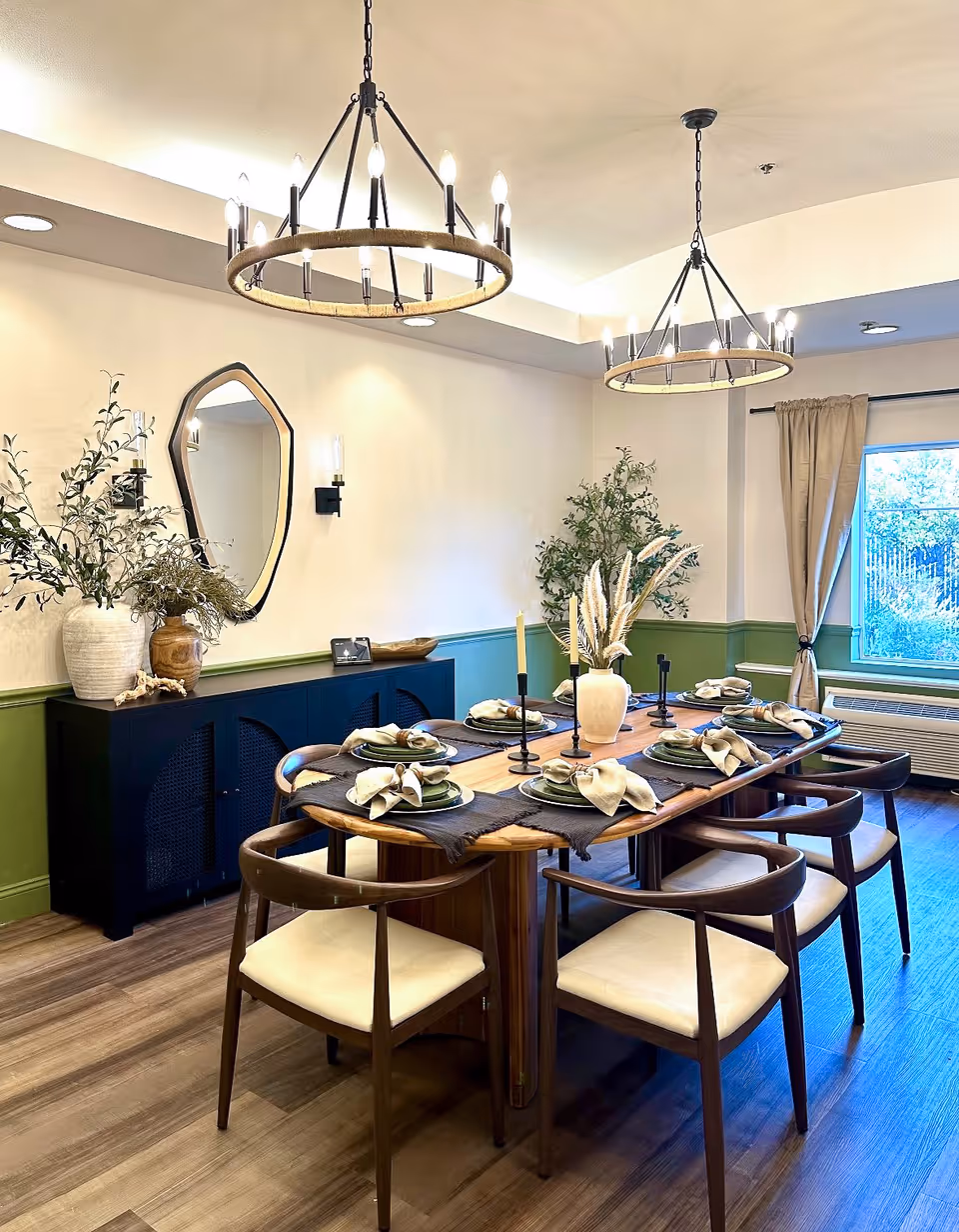 A well-lit dining room with a wooden dining table set for six, featuring plates, napkins, and candles. The room has two circular chandeliers hanging from the ceiling, a large window with beige curtains, a black sideboard with decorative vases and plants, and a uniquely shaped mirror on the wall. The lower half of the walls is painted green, and the floor is wooden.