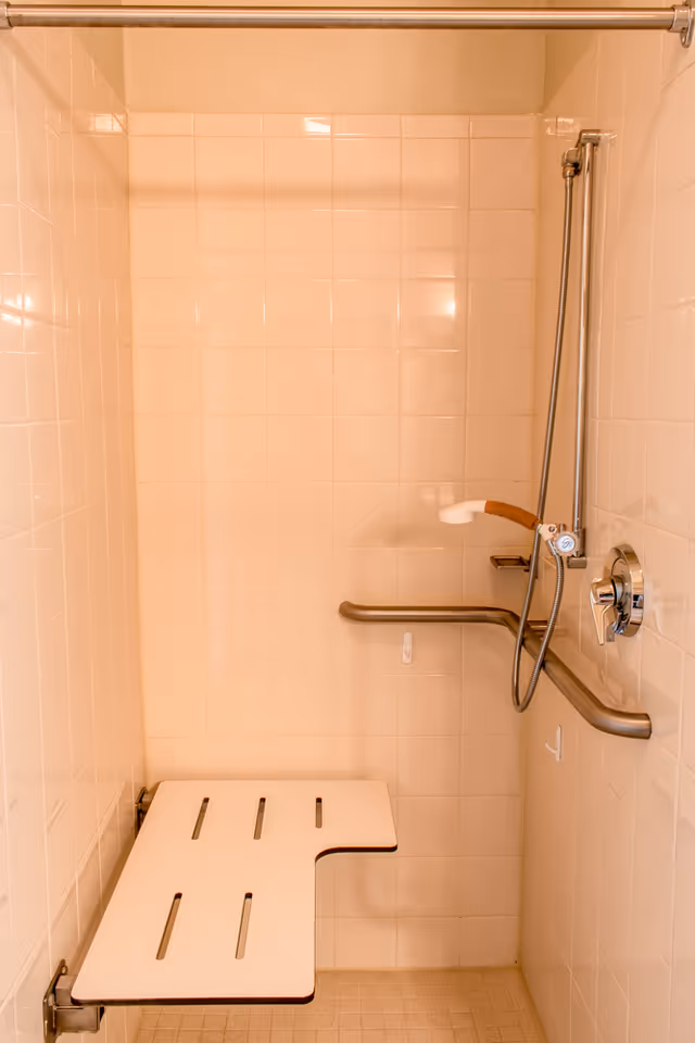 A tiled shower area with a foldable white shower seat mounted on the wall, a handheld showerhead attached to a vertical bar, and a horizontal grab bar for support. The shower has a metal curtain rod at the top.