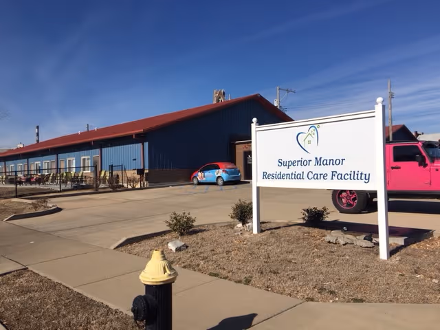 Exterior view of Superior Manor Residential Care Facility showing a long blue building with a red roof, a parking area with a blue car and a pink vehicle, a sidewalk, a fire hydrant, and a white sign with the facility's name.