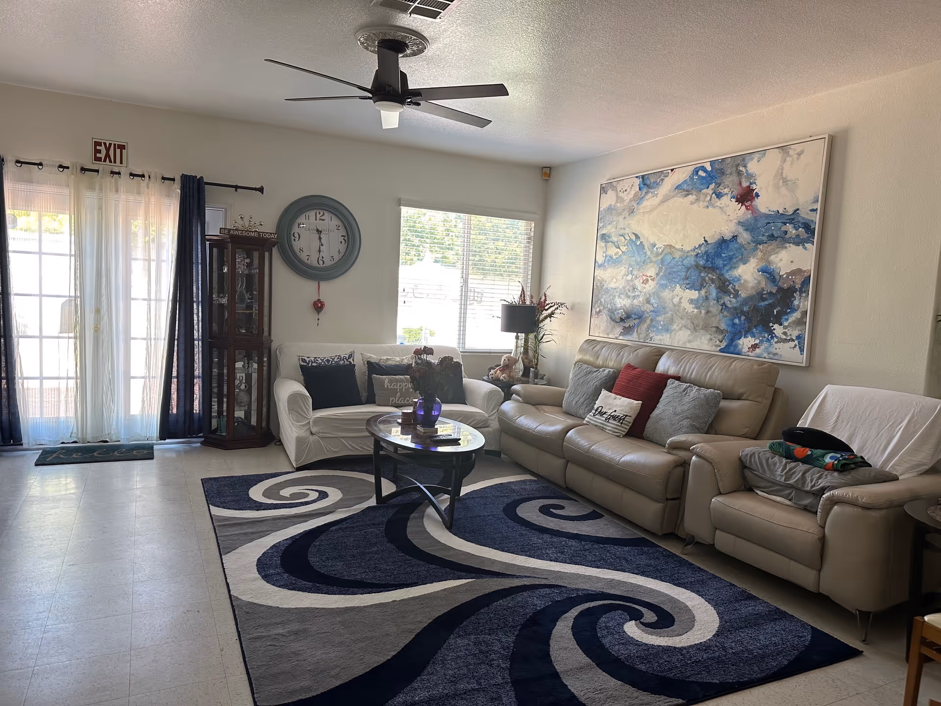 A cozy living room with a large abstract blue and white painting on the wall above a beige leather sofa and armchair. There is a white loveseat with decorative pillows, a round coffee table with a purple vase, and a patterned blue and gray rug on the floor. A large clock hangs on the wall near a window with blinds, and an exit door with sheer and dark curtains is visible on the left side.