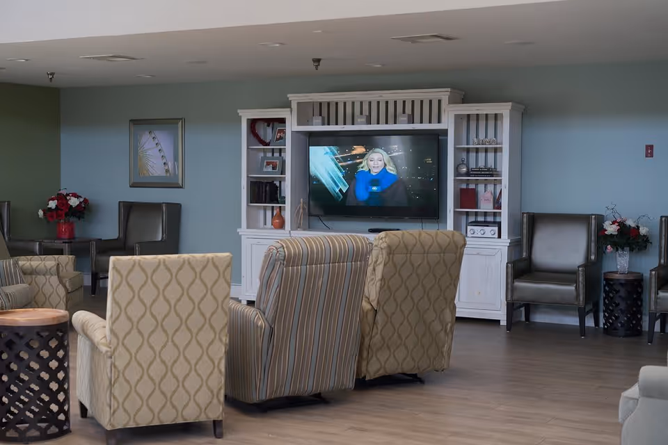 A cozy living room area in a senior living facility with several upholstered armchairs arranged facing a white entertainment center with a flat-screen TV. The TV displays a news anchor. The walls are painted light blue and decorated with framed artwork and flower arrangements on side tables.