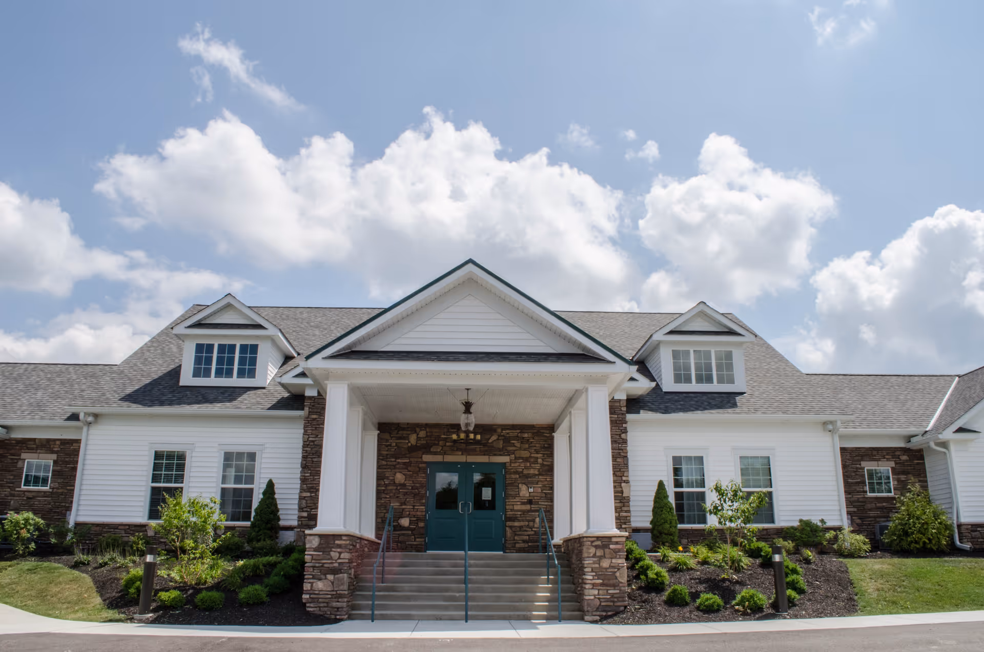 Front exterior view of Meadow Falls Of Chagrin Valley building with white siding, stone accents, green double doors, and a covered entrance with steps leading up. The sky is partly cloudy.
