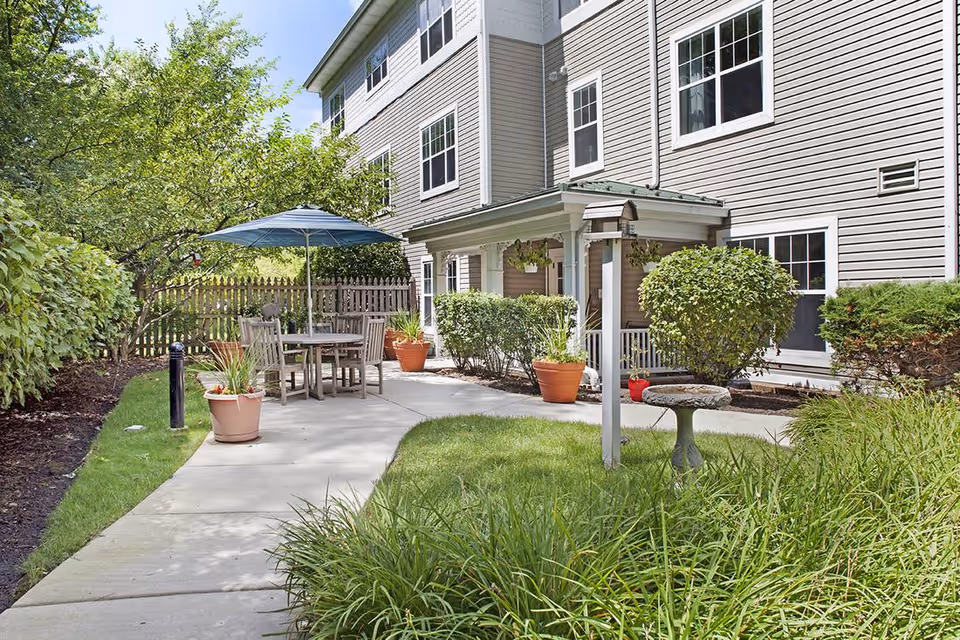 Outdoor patio area at Sunrise of Willowbrook featuring a concrete walkway, wooden table with chairs under a blue umbrella, potted plants, trimmed bushes, a birdbath, and a three-story building with multiple windows in the background.