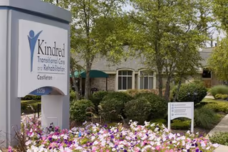 Exterior view of Kindred Transitional Care and Rehabilitation Castleton building with a sign in the foreground surrounded by colorful flowers and trees.