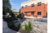 Outdoor view of a senior living facility named Spring Creek featuring a landscaped garden with a stone pathway, plants, and a two-story brick building in the background under a clear sky.