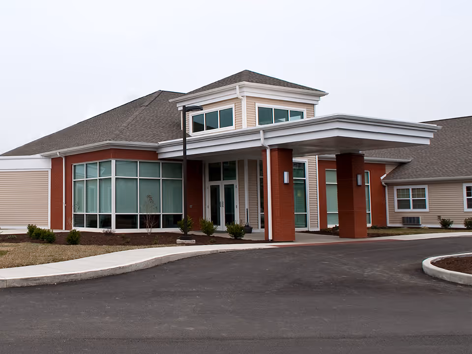 Exterior view of BridgePort Healthcare Center showing the main entrance with a covered drop-off area supported by brick columns, large windows, and a paved driveway with landscaped areas.