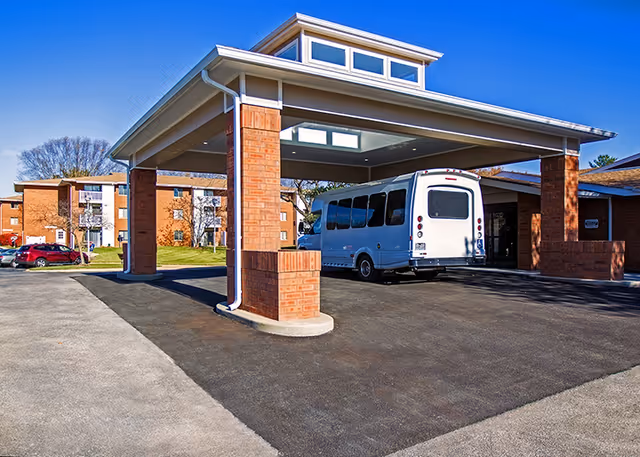 Covered entrance area of a senior living facility with a white shuttle bus parked underneath. The building features brick pillars and a peaked roof with windows. In the background, there are multi-story brick residential buildings and a few parked cars on a sunny day.
