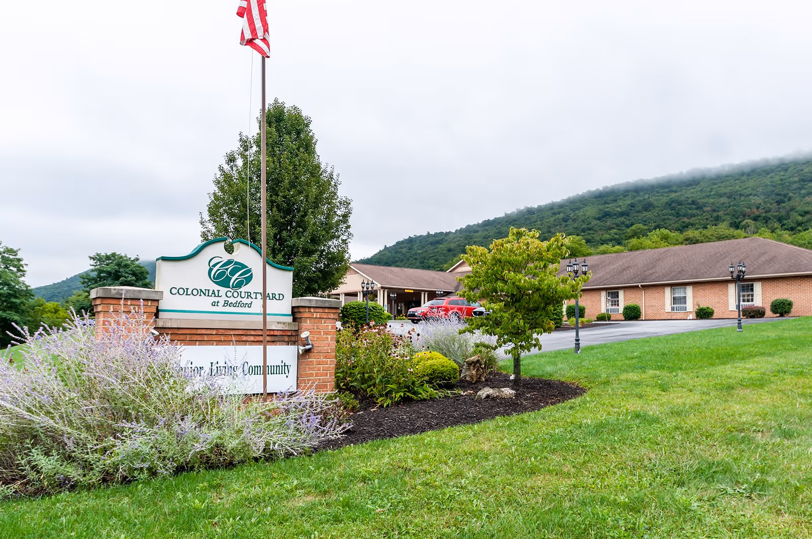 Exterior view of Colonial Courtyard at Bedford senior living community with a brick sign, an American flag on a pole, landscaped bushes and flowers, a green lawn, and a building with a brown roof in the background under a cloudy sky and hills.