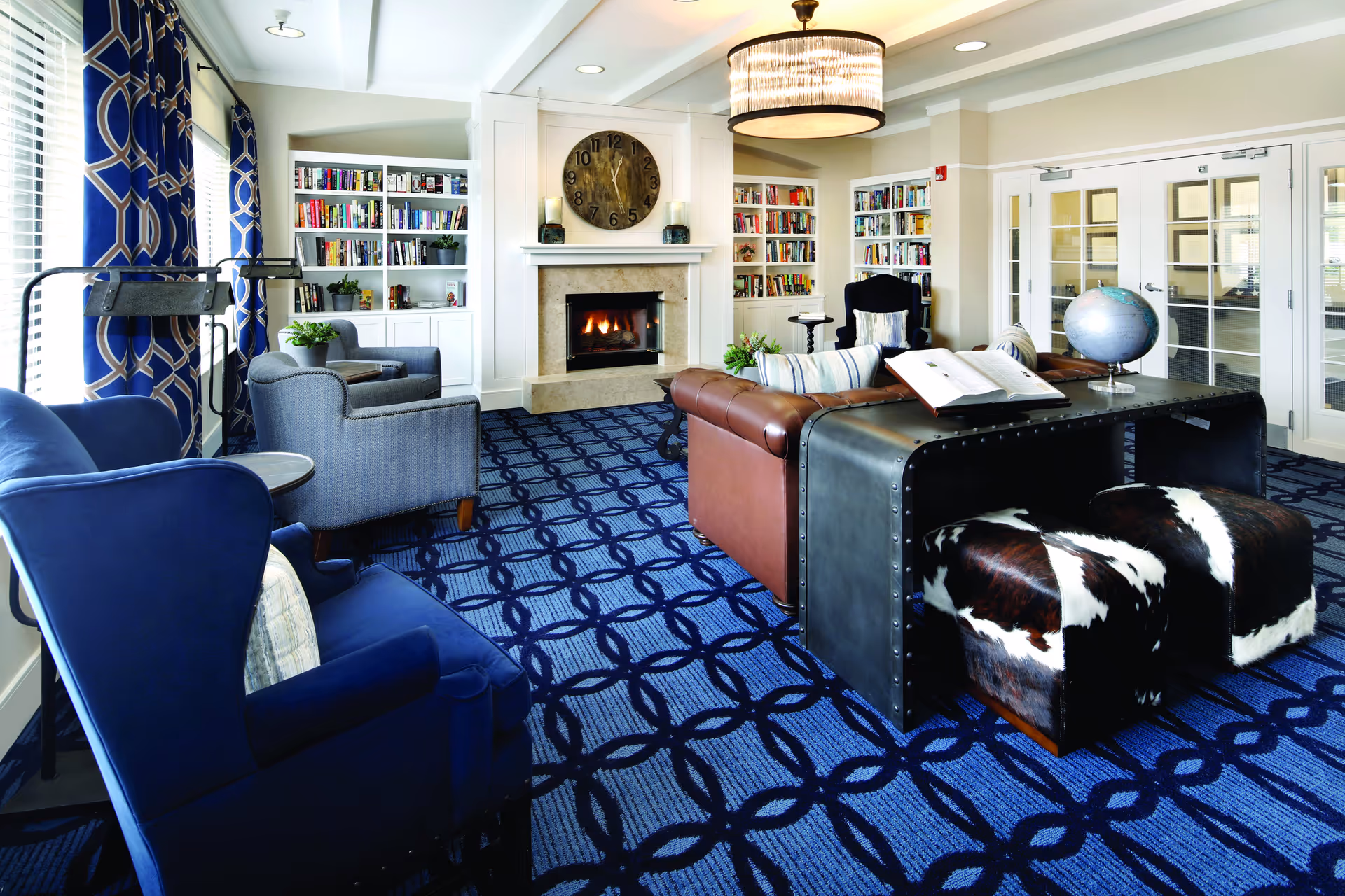 A cozy living room with a blue patterned carpet, blue and gray armchairs, a brown leather sofa, and a black metal desk with two cowhide stools. The room features a fireplace with a large clock above it, built-in white bookshelves filled with books, and a large window with blue patterned curtains. A globe and an open book are on the desk, and there is a modern chandelier hanging from the ceiling.