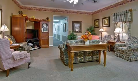 A cozy living room area in a senior living facility with patterned armchairs, a wooden coffee table decorated with a floral centerpiece, a wooden entertainment center with a TV, and lamps providing warm lighting. The room has beige walls with framed artwork and a window with plaid curtains. A hallway is visible in the background.