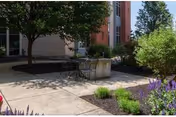 Outdoor patio area with a small round table and two chairs surrounded by trees, shrubs, and flowering plants near a building.