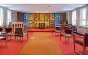 Interior view of a chapel or meditation room with a red carpeted floor and several chairs arranged in rows facing a colorful stained glass window at the front. The room has white walls and windows on both sides allowing natural light to enter.