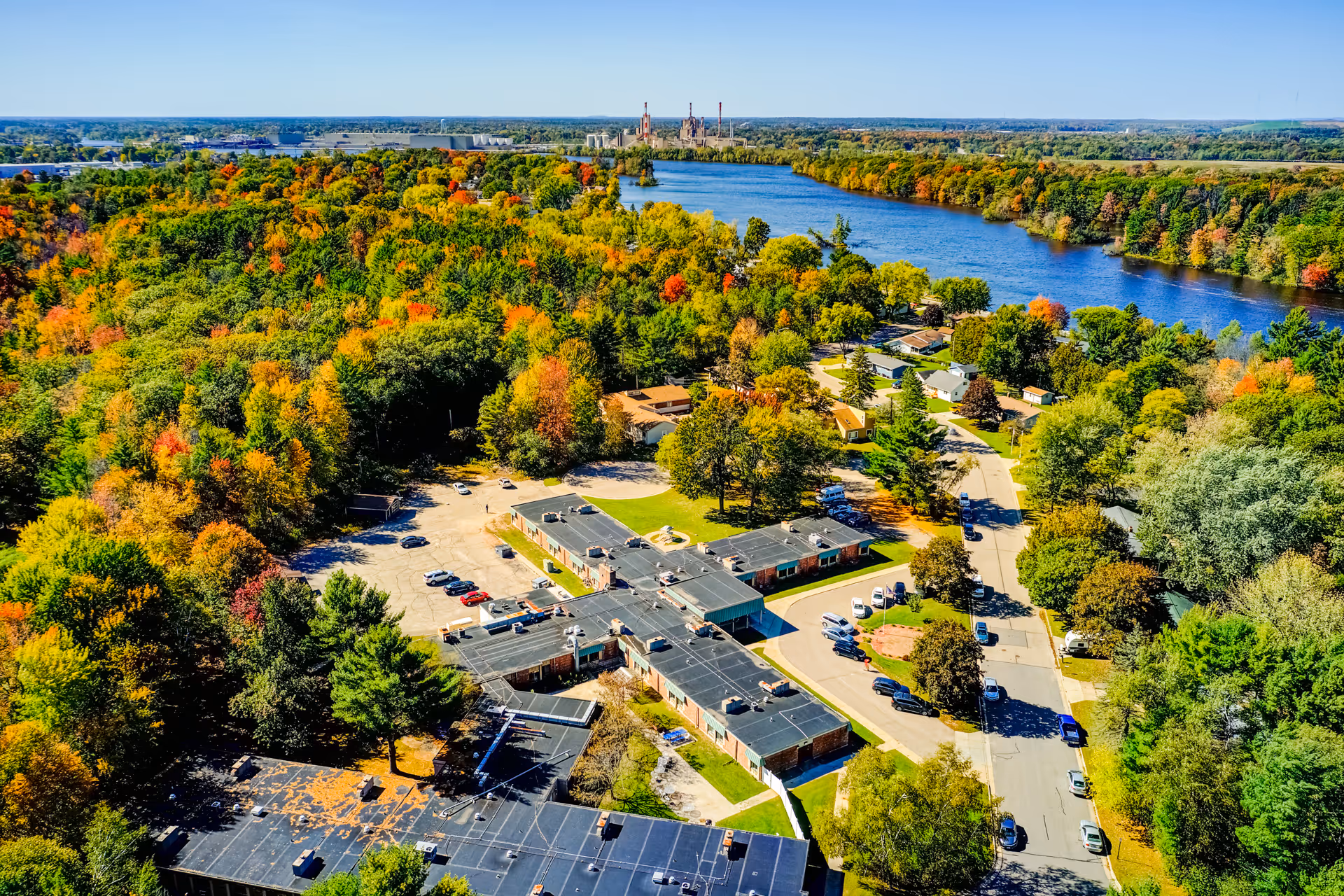 Aerial view of Edenbrook Wisconsin Rapids facility surrounded by dense trees with autumn foliage near a river, showing the building complex, parking lots, and nearby residential area under a clear blue sky.
