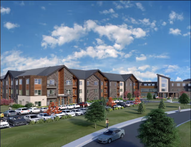 Exterior view of a large, modern three-story senior living facility named Astral at Auburn, with a spacious parking lot filled with cars, landscaped green areas with trees and shrubs, and a partly cloudy blue sky above.