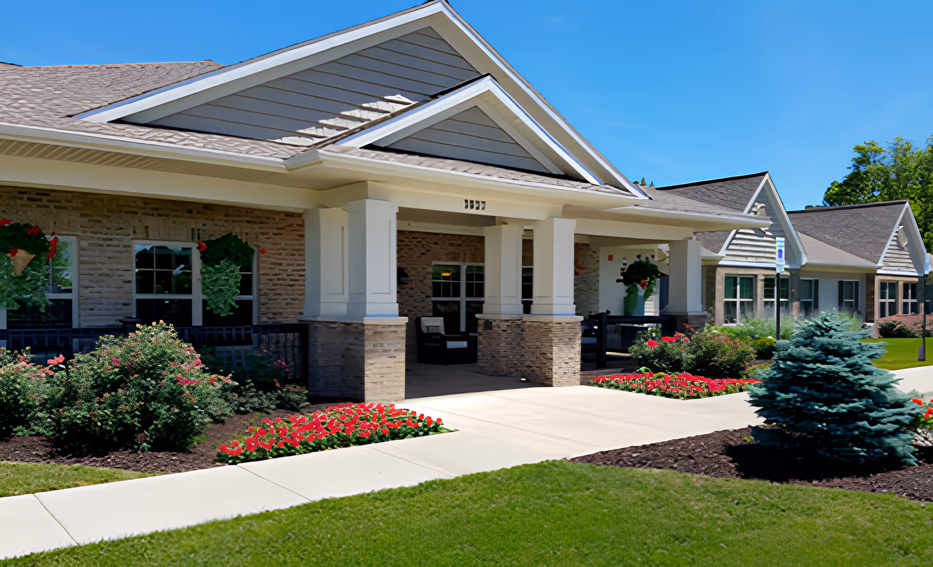Exterior view of a single-story retirement living facility with a covered entrance supported by white columns. The building has a combination of brick and siding with a gabled roof. The landscaped front area features green grass, bushes, red flowers, and a small evergreen tree under a clear blue sky.